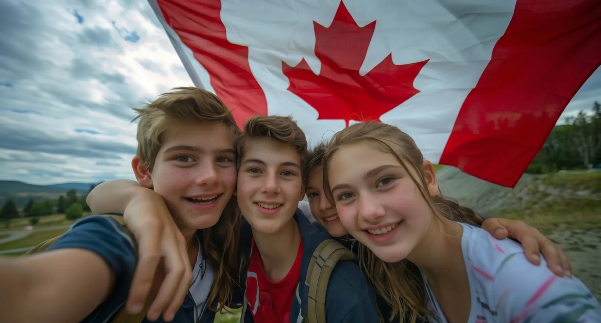 Un grupo de cuatro estudiantes tomándose un selfie muy sonrientes y abrazados, con una gran bandera de Canadá ondeando directamente detrás de ellos en un paisaje natural al aire libre en su año escolar en Canadá.