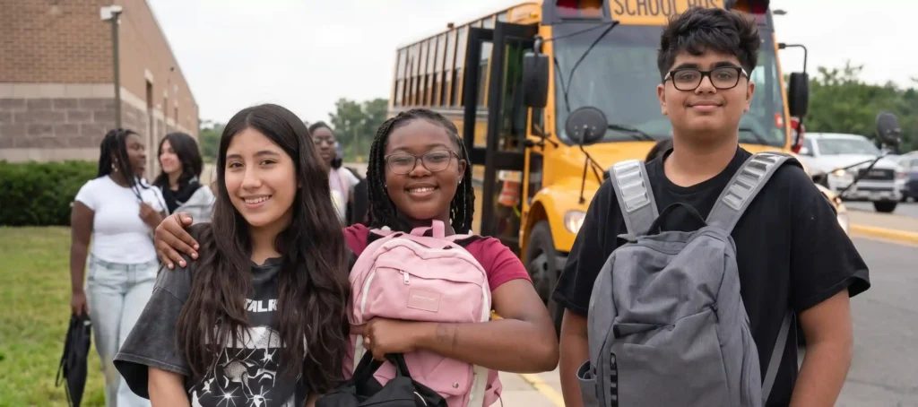 Un grupo de tres adolescentes sonriendo y sosteniendo mochilas frente a un autobús escolar amarillo con las puertas abiertas, afuera de un edificio escolar, ilustrando la experiencia de intercambio de estudiantes en Estados Unidos gracias al Visado F1 Estados Unidos