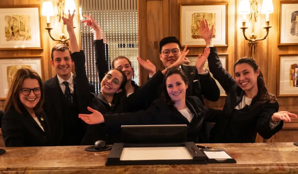 Grupo de siete empleados de recepción con uniformes formales oscuros celebrando y levantando los brazos detrás de un mostrador de madera clásico.