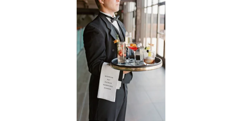 Camarero profesional con smoking sosteniendo una bandeja de cócteles elegantes, representando el puesto de Servidor para el programa Work and Travel USA en hostelería.