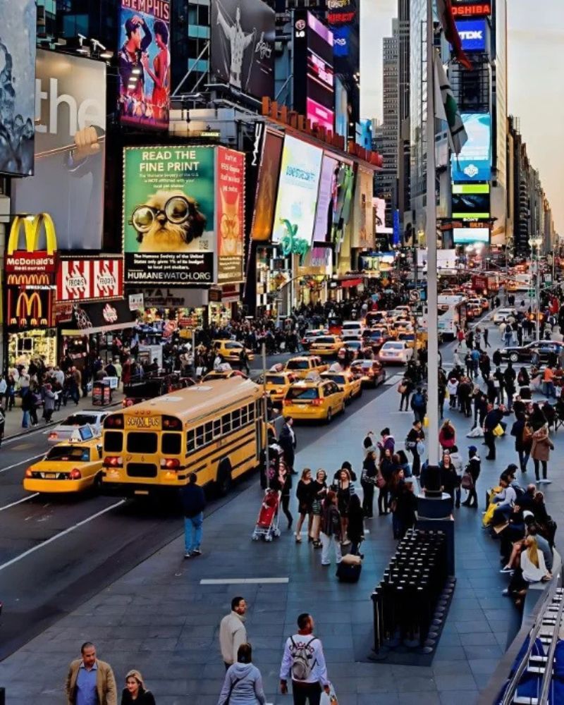 Multitud de peatones, taxis amarillos y un autobús escolar bajo las famosas luces y pantallas gigantes de Times Square en Nueva York, un destino imprescindible al viajar por Estados Unidos.