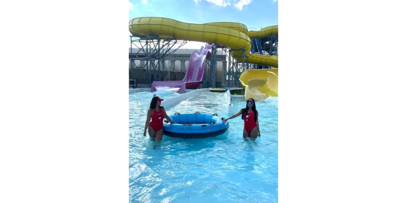 Dos jóvenes socorristas con uniforme rojo y viseras posan en una piscina de un parque acuático en Estados Unidos sujetando un flotador azul gigante, con grandes toboganes amarillos y morados al fondo. Un ejemplo de empleo popular para participantes del programa Work and Travel USA.