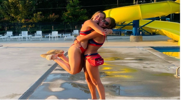 Dos socorristas con bañadores rojos y azules abrazándose de forma divertida en el borde de una piscina exterior con un tobogán amarillo de fondo.