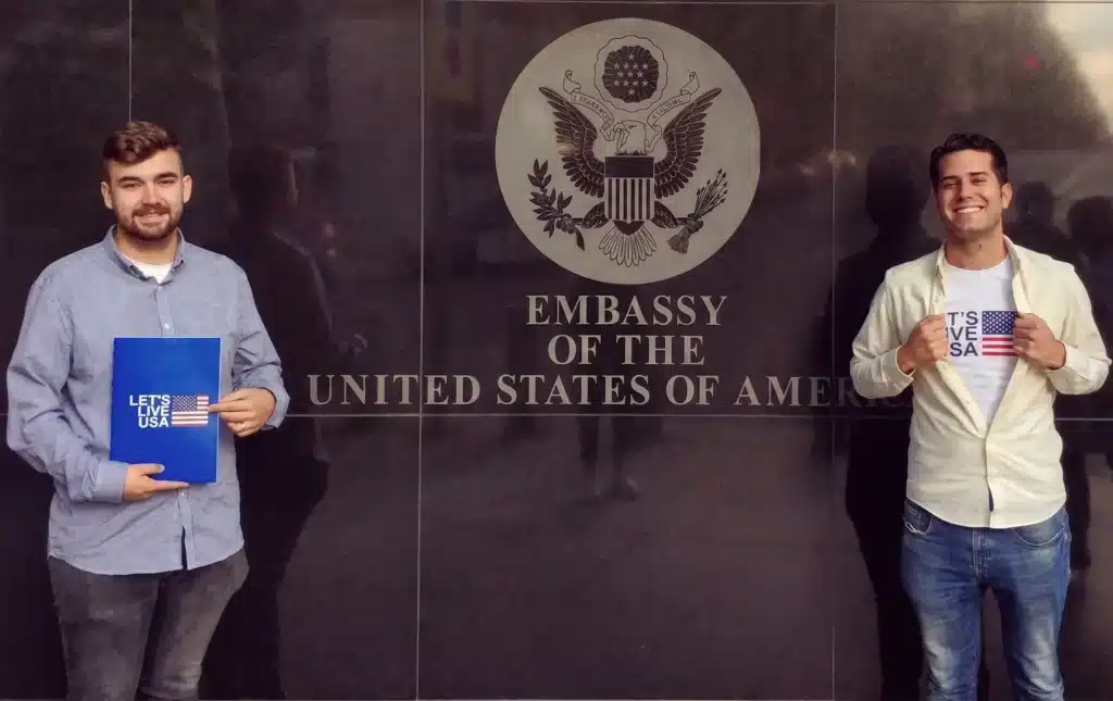 Dos jóvenes sonrientes posan frente a la fachada de la Embajada de los Estados Unidos. Uno sostiene una carpeta azul y el otro muestra una camiseta con el logo "Let's Live USA" y la bandera estadounidense.