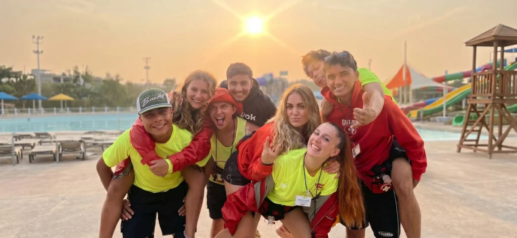 Grupo Socorristas Work and Travel USA Parque Acuatico Grupo de jóvenes del programa Work & Travel USA en un parque acuático durante el atardecer. Los participantes visten uniformes de socorrista con camisetas amarillas y chaquetas rojas, posando felices y abrazados, con algunos compañeros a caballito, reflejando el excelente ambiente laboral y la oportunidad de viajar barato por Estados Unidos