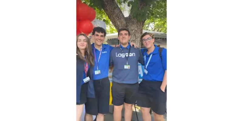 Grupo de cuatro jóvenes participantes del programa Work and Travel USA posando felices y abrazados bajo un árbol. Visten uniformes azules con acreditaciones para su trabajo como Ride Operators (operadores de atracciones) en un parque de diversiones.